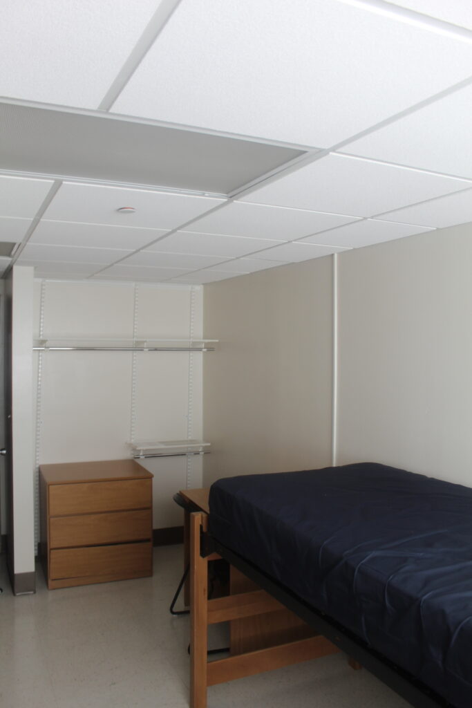 An undecorated bedroom with a twin XL mattress, dresser and desk with a chair