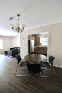 The dining area that features a round dining table and four black metal chairs around it. It features an uncentered chandelier and a view of the modern furniture in the living room and a glimpse of the kitchen in the back of the photo