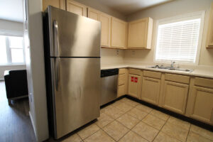 A view of the kitchen featured light toned wood cabinets, a double stainless steel sink, a window above the sink and a stainless steel fridge and dishwasher