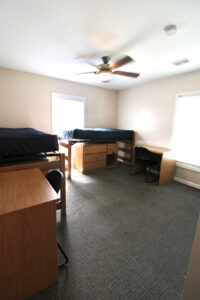 A undecorated bedroom that features two twin XL beds, dressers underneath the lofted beds and desks on either side with black chairs