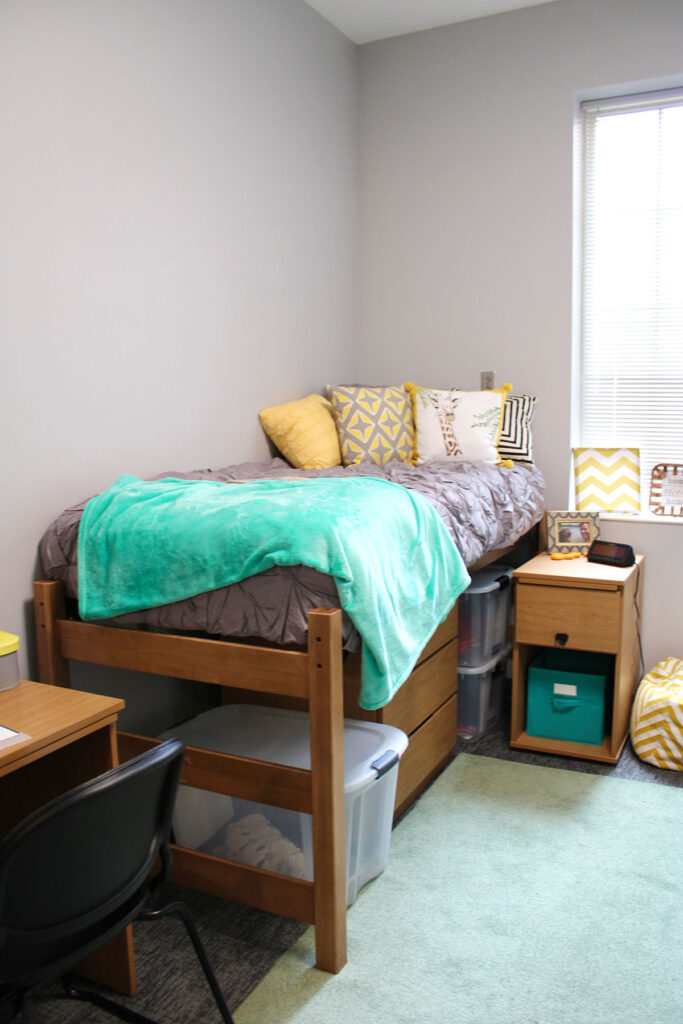 A furnished bedroom in Magnolia Hall that features a mint green rug, yellow pillows and a gray comforter