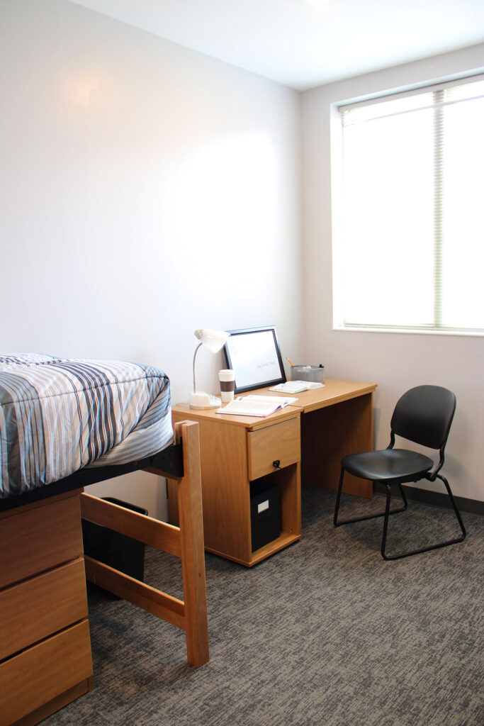 A furnished bedroom in Magnolia Hall that focuses on the desk in the corer of the room with a black chair, a desk lamp and a coffee cup