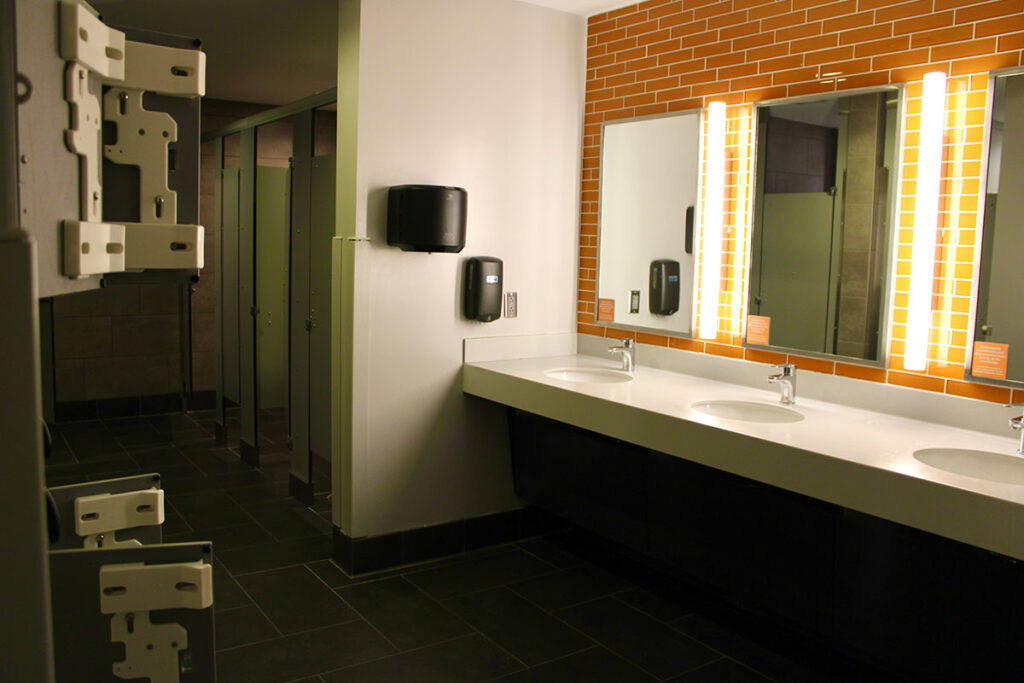 The bathroom in Magnolia Hall that features horizontal orange tiles behind the three mirrors in the floating vanity with stalls to the left back of the room