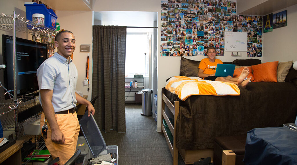 Two students in a decorated dorm. One student is working on their laptop in one bed with photos covering the wall while the other continues working on unpacking. Both are smiling at the camera.