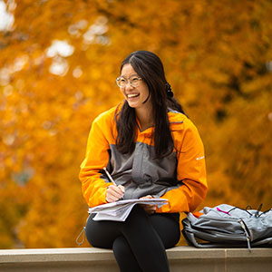 Student sitting with a notebook laughing. Wearing an orange and grey zipup and fall leaves are in the background