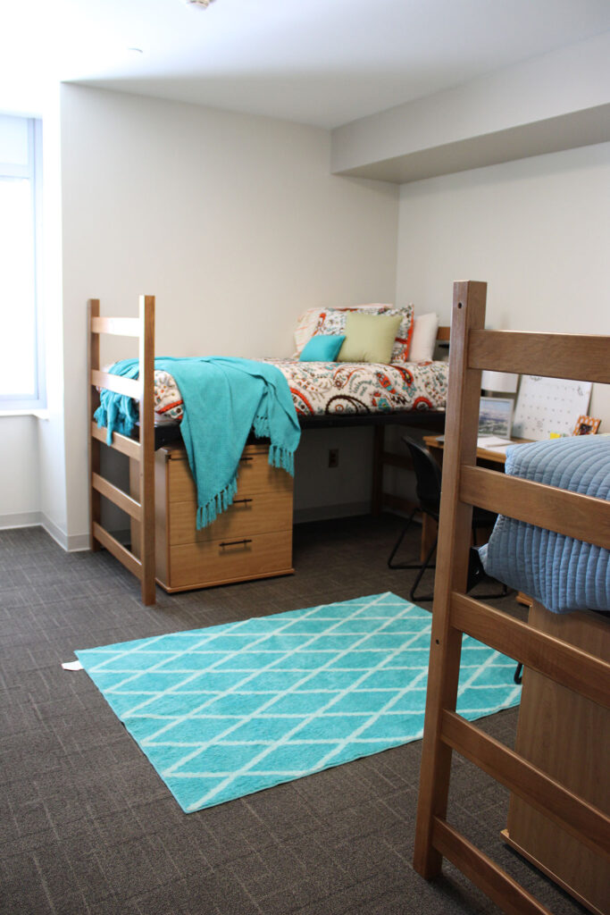 A decorated dorm room with lofted beds that have a dresser underneath each one, two desks in between the beds and a turquoise rug in the middle of the room