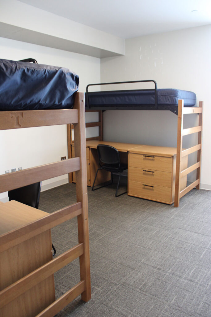 An undecorated dorm room with two beds that are lofted and a desk and black chair under each desk