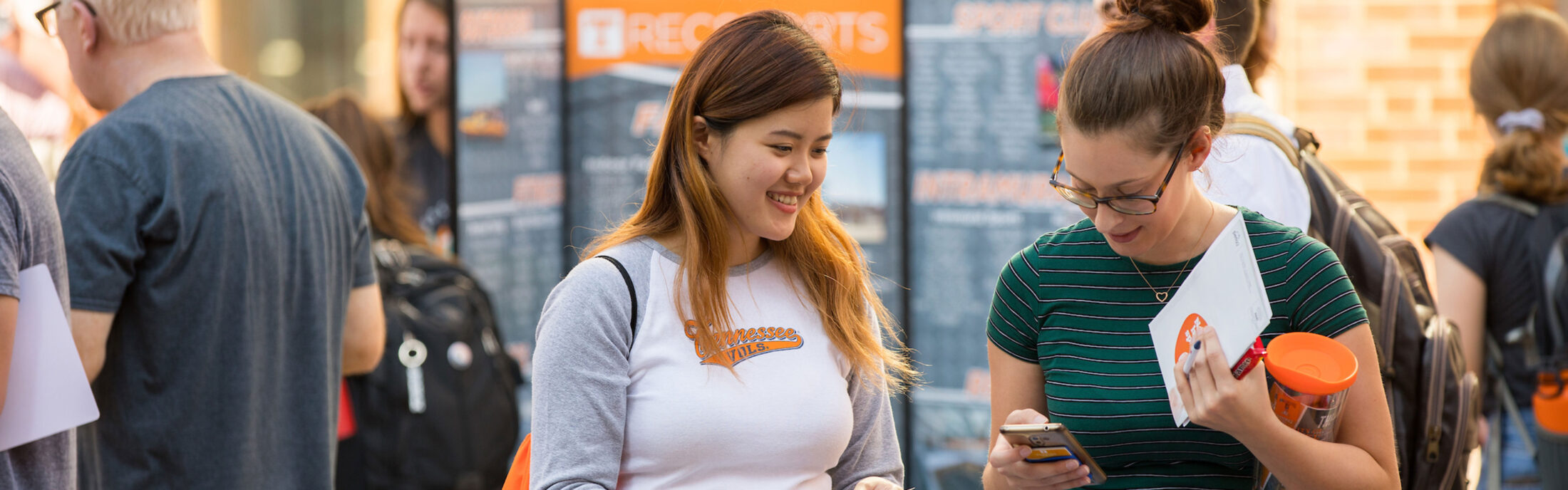 Two students attend the engagement fair at the Student Union Plaza. One student is viewing a phone.