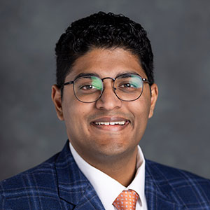 Staff headshot of Sohan Daniel wearing a blue suit with an orange tie, sitting in front of a dark gray backdrop.