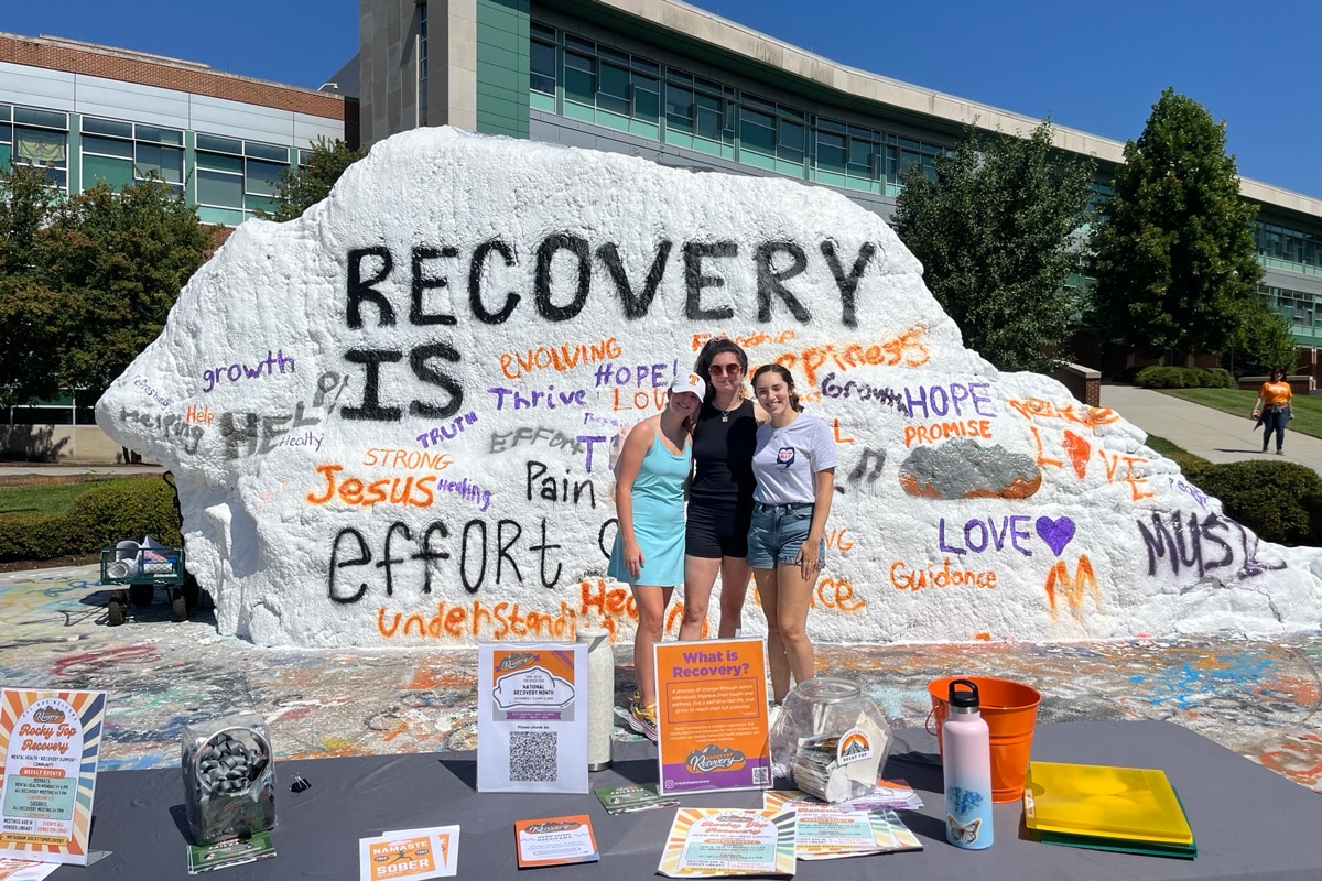 two individuals standing in front of the rock at UT with the words "Recovery Is"