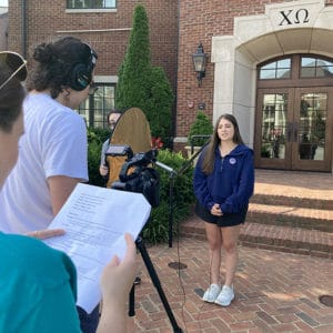 two students interviewing a female student in front of the Chi Omega sorority house