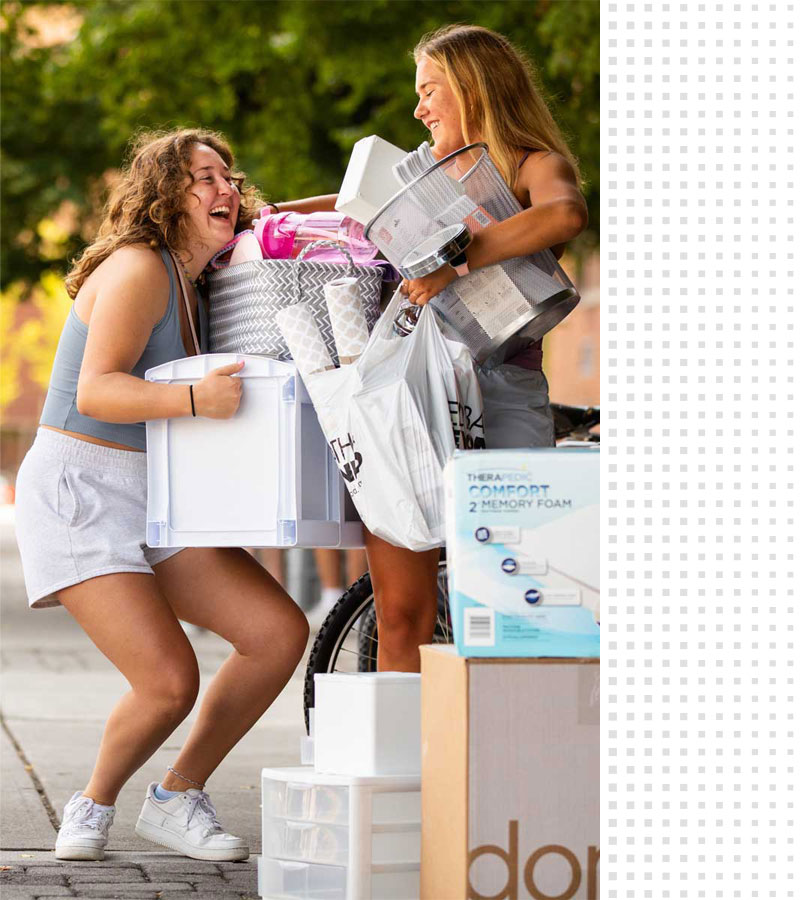 Two students laugh with arms full of home goods as they move into their residence hall