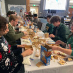 Picture of many individuals at a gingerbread house making event making gingerbread houses down a long table.