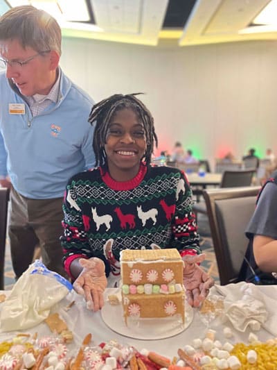 Picture of an individual posing with the finished product of her gingerbread house making, wearing a Christmas sweater.