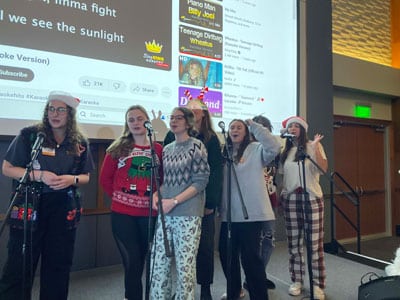 7 individuals singing karaoke on stage all wearing some sort of Christmas pajamas. Two are wearing Santa hats.