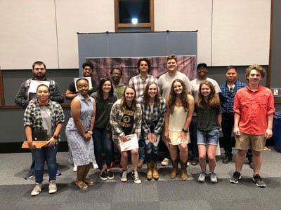 Picture of 15 individuals in front of a sign holding various items.