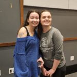 Picture of two individuals, one on the left has curly hair and is in a blue sweater holding a water cup and the one on the right is in a grey shirt an hair is in an updo.