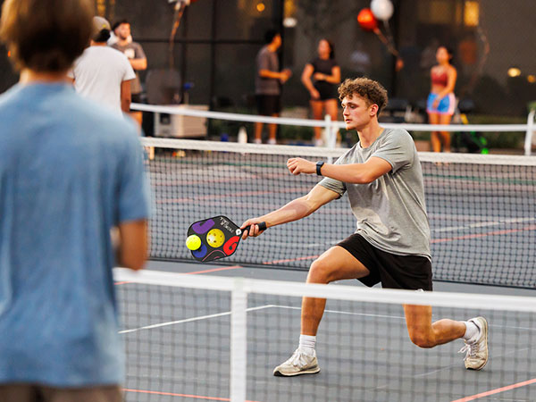 A male student leans in to swing at the ball with his pickle ball paddle while his opponent anticipates the hit on the other side of the net.
