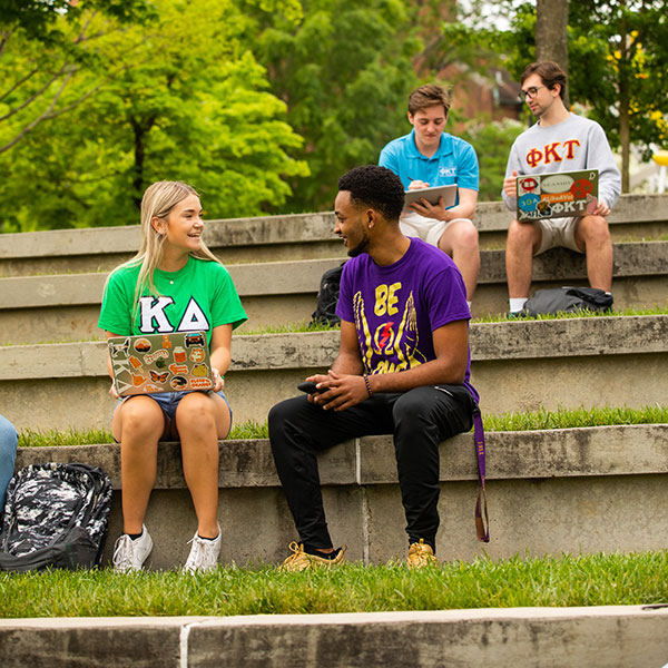 Students wearing their Greek letters converse outside on grass steps in the Humanities Amphitheater.