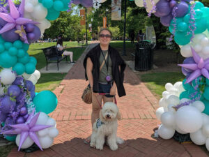 Kori Turney and her service dog pose for a photo underneath a purple and blue balloon arch on a red brick walkway in Circle Park.
