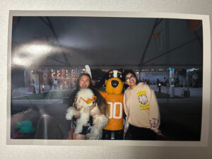 A printed photo sitting on a tabletop depicts Kori Turney lifts up her service dog to pose for a photo with Smokey the mascot and a friend.